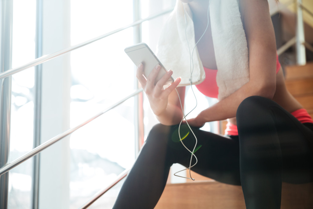 Closeup of sportswoman with white towel sitting and using smartphone with earphones in gym
